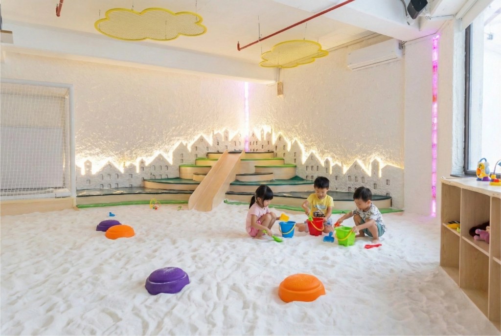 Three young children happily playing with colorful sand toys, buckets, and shovels in the deep white granular salt floor of the therapeutic kids' salt room at Filifu Cafe, featuring a wooden slide and illuminated city wall decor.