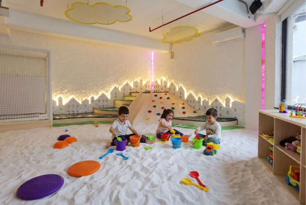 Filifu Cafe'nin İstanbul Boğazı ve Marmara Denizi manzaralı benzersiz çocuk tuz odası oyun alanı. Beyaz granül tuz kaplı zeminde renkli kovalarla oynayan üç çocuk, ahşap kaydırak, tırmanma duvarı, ışıklı şehir silüeti ve bulut ışıklar görülüyor.