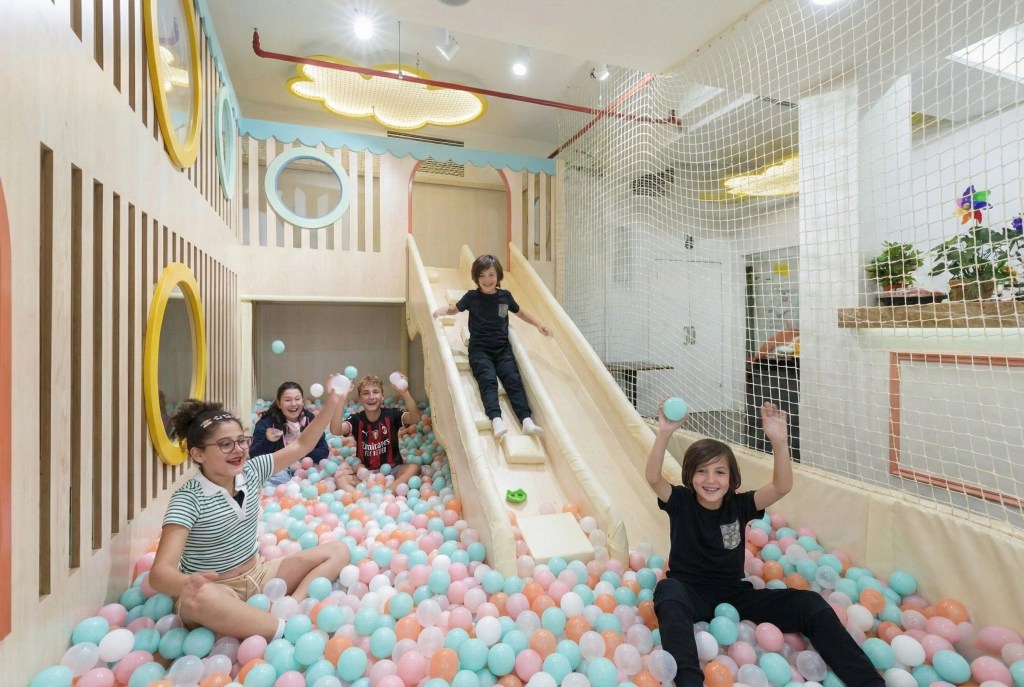 Filifu Cafe 梦幻波波池内，几个快乐的孩子正从定制的木制滑梯滑下，在充满马卡龙色系（粉色、蓝色、白色）海洋球的池中尽情嬉戏互动。