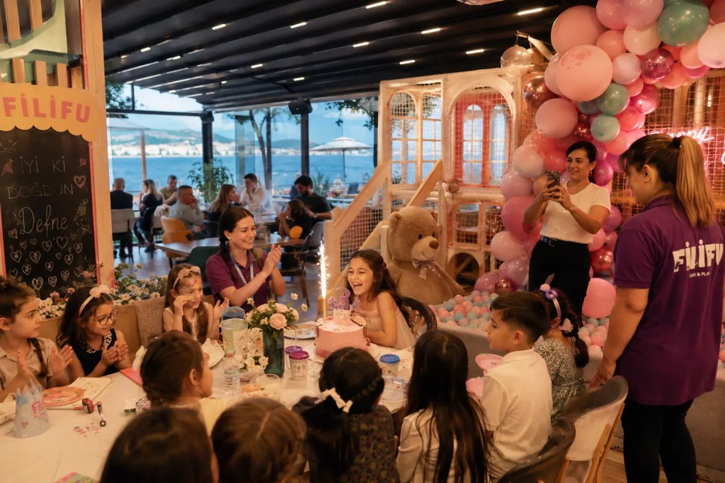 Filifu Cafe N’ Play’de düzenlenen konsept çocuk doğum günü partisinde çocuklar pasta keserken, oyun ablaları etkinliğe eşlik ediyor ve arka planda deniz manzarası ile oyun alanı görülüyor.