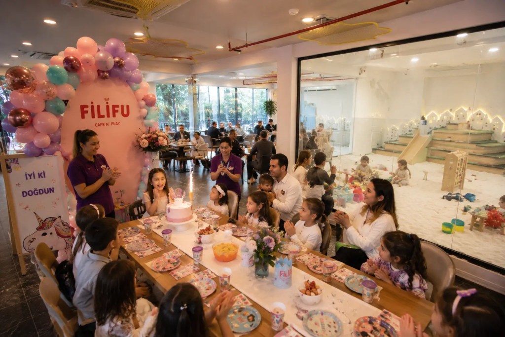 Filifu Cafe N’ Play’de düzenlenen çocuk doğum günü kutlamasında çocuklar pasta kesimi için toplanırken, arka planda camla çevrili tuz odası ve aile oturma alanları görülüyor.