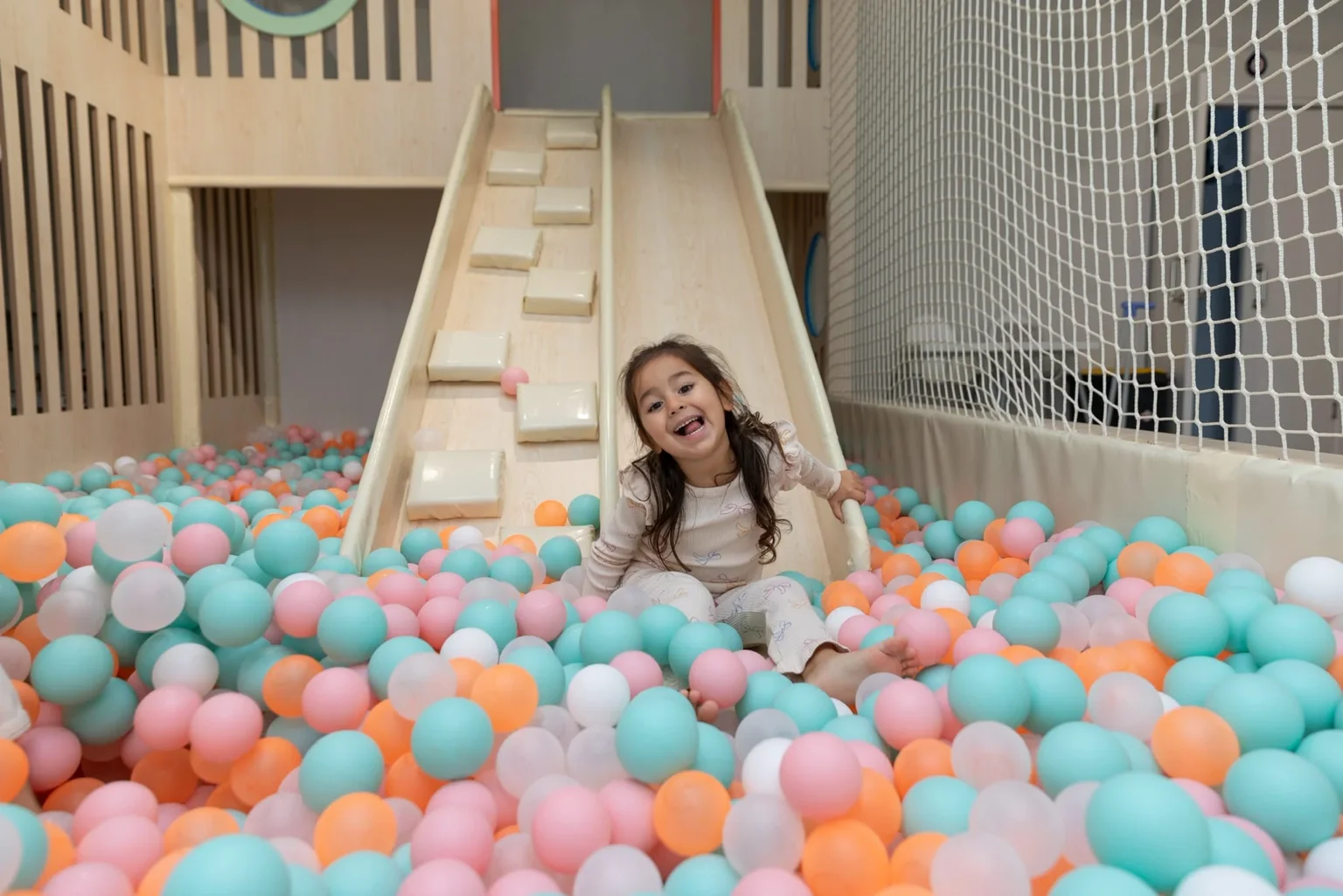 A child seated among pastel balls beside a pale wooden slide and white safety net