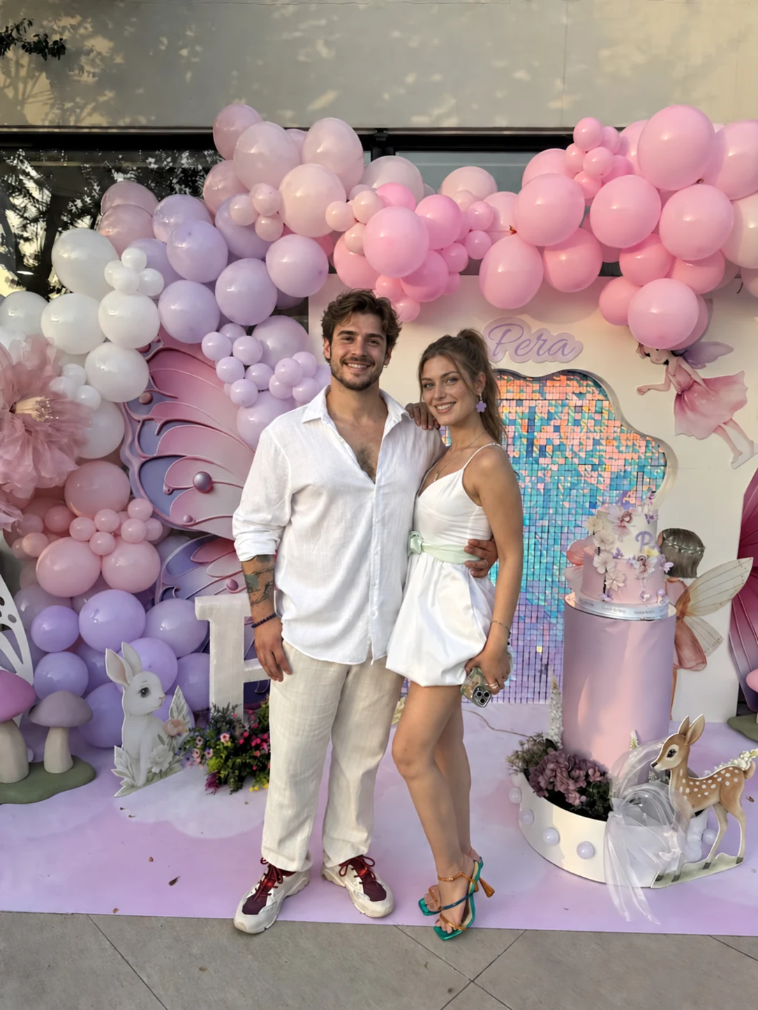Filifu'da pembe ve lila balonlarla süslenmiş, peri temalı pasta köşesinin önünde duran iki yetişkin.