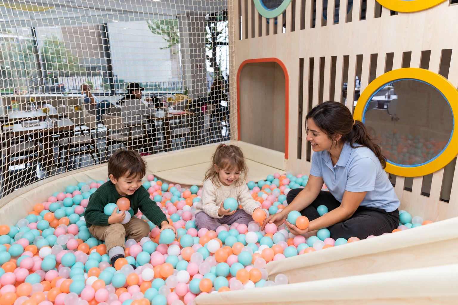 Two children holding pastel balls beside a play assistant in Filifu’s wooden ball-pit area