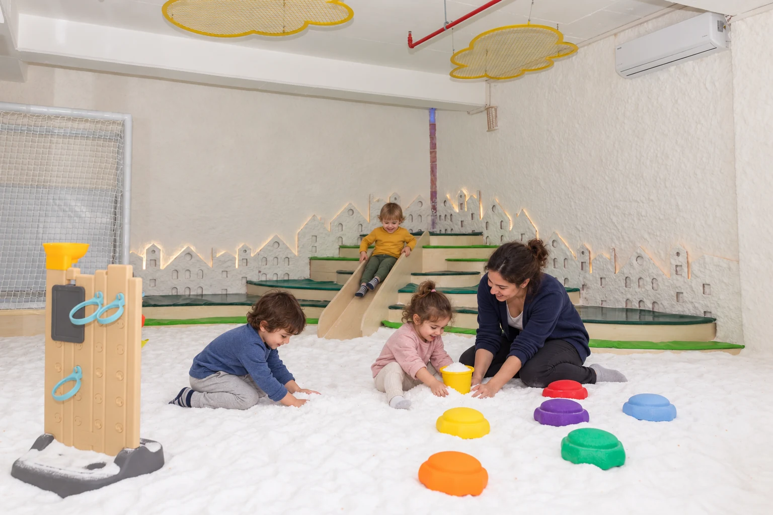 Three children and a play assistant exploring Filifu’s salt room with a yellow bucket and a wooden slide behind them